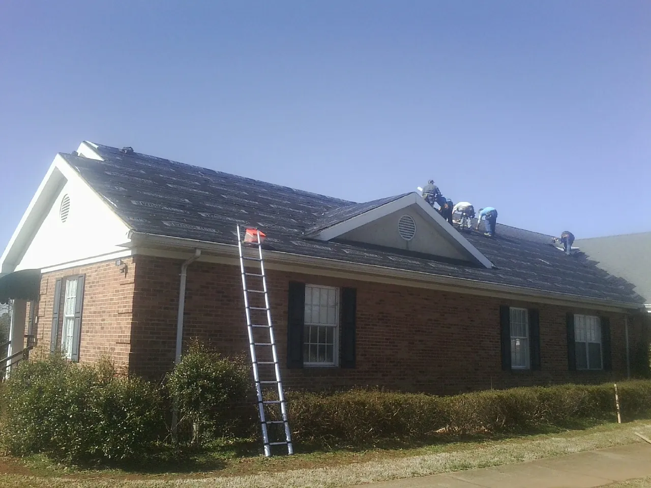 Crew tearing shingles off a brick ranch roof with a ladder set — Emergency Roof Repair work by our crew in Camby