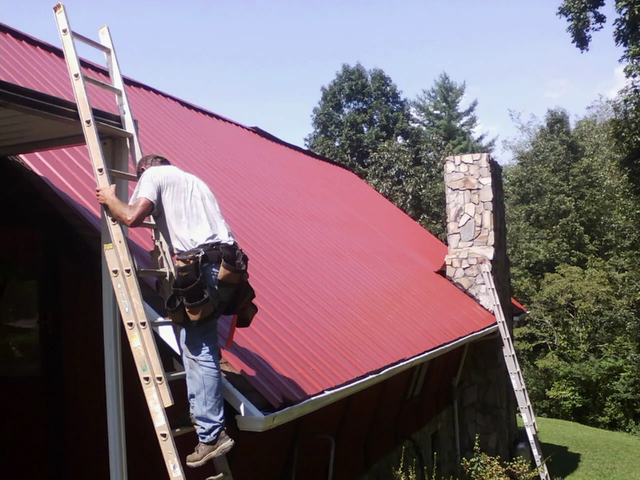 Roofer on a ladder beside a red metal roof and stone chimney — Soffit & Fascia Repair by Roofing Brothers in Camby